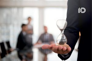 A person holds an hourglass with a business meeting in the background, demonstrating The Time and Material Pricing Model.