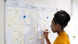 A young woman with long hair gestures while explaining Kanban, a visual project management method using cards on a board.